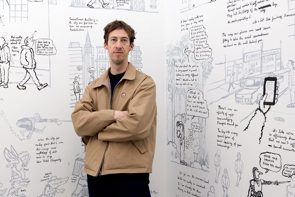 Artist Jean Jullien poses with his arms crossed in front of his artwork, a black and white doodle mural on a white wall in an art gallery. 
