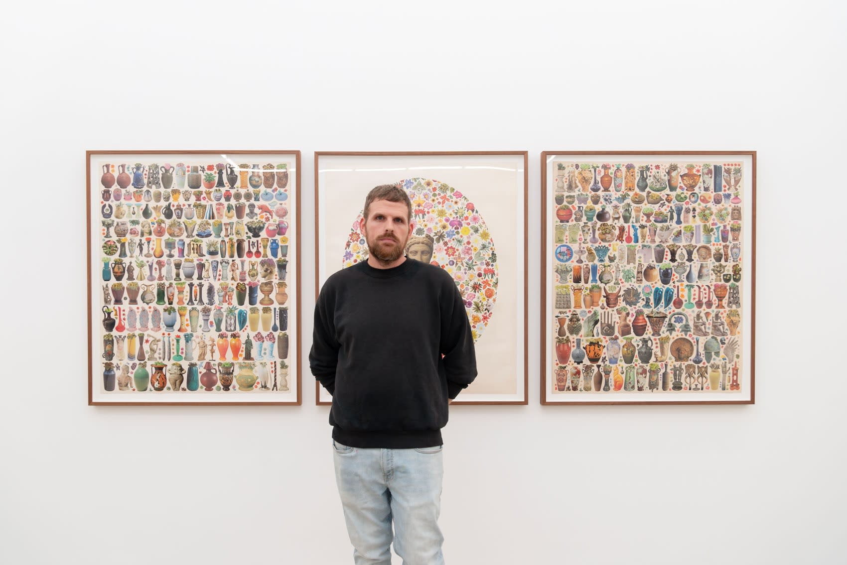 Matthew Craven, a tall man in a black sweater, stands in front of three of his large collage works on paper, framed, in a white cube gallery