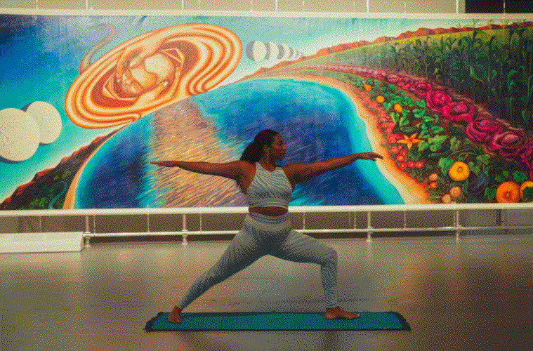 A person holds a Warrior II yoga pose on a blue mat in front of a colorful mural with an abstract, cosmic landscape and flowers. The room is brightly lit, enhancing the tranquil ambience.