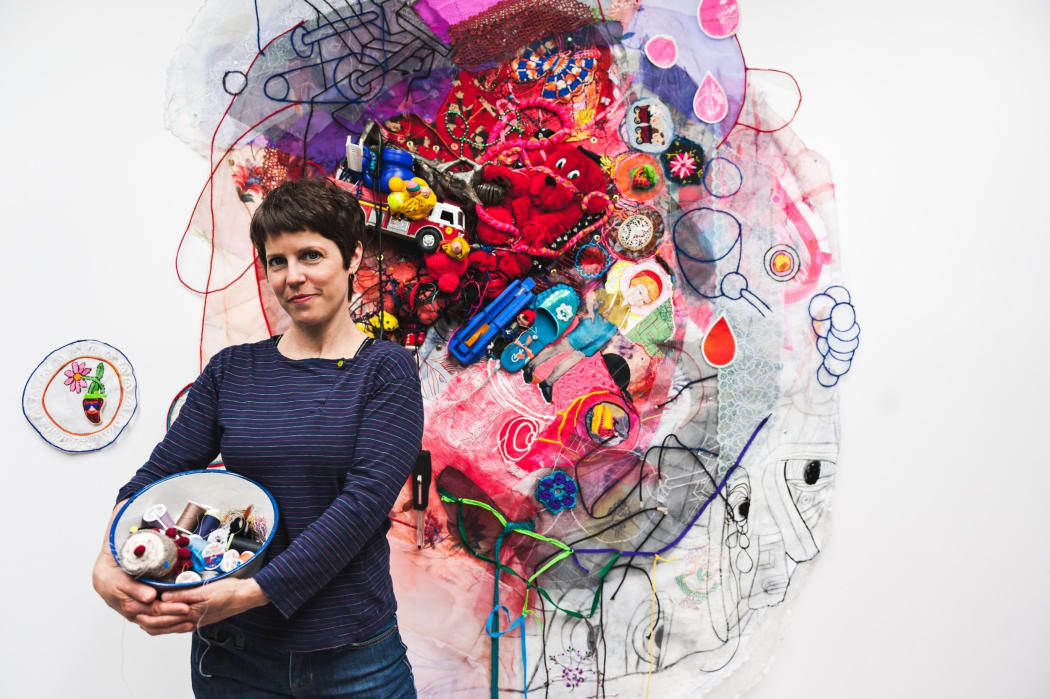 Artist Julie Peppito in a striped blue shirt cradling a bowl of sewing supplies to the left in front of one of her own multi-color assemblage installations.