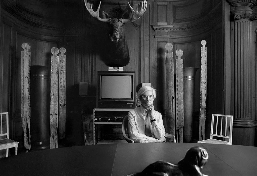 Andy Warhol seated in the conference room at the Factory with vigango, VCR Television, and moose head taxidermy mounted on the wall, photographed by Robert Levin, 1981. Vigango have been celebrated and collected by artists and collectors around the world, including Warhol.