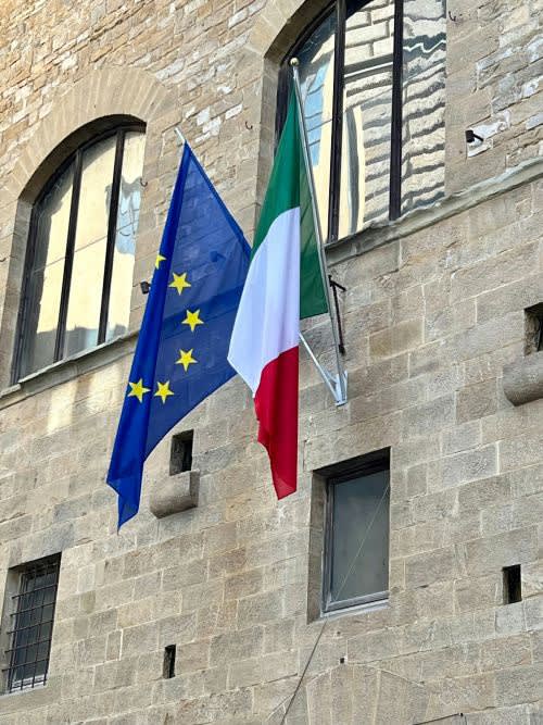 An Italian flag and European Union flag hanging side by side on an old Italian building facade