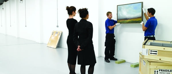 Photograph of two men installing a painting on a wall at an art gallery. Two women observe as it is installed.