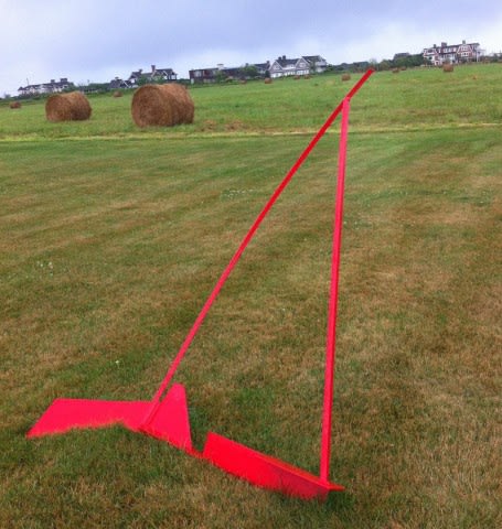 This is a photograph of an art piece that is sitting on a field with the flat surface having an angle. The art piece's placement as well as having a bright red color makes it stand out compared to the houses and hay rolls in the distance.