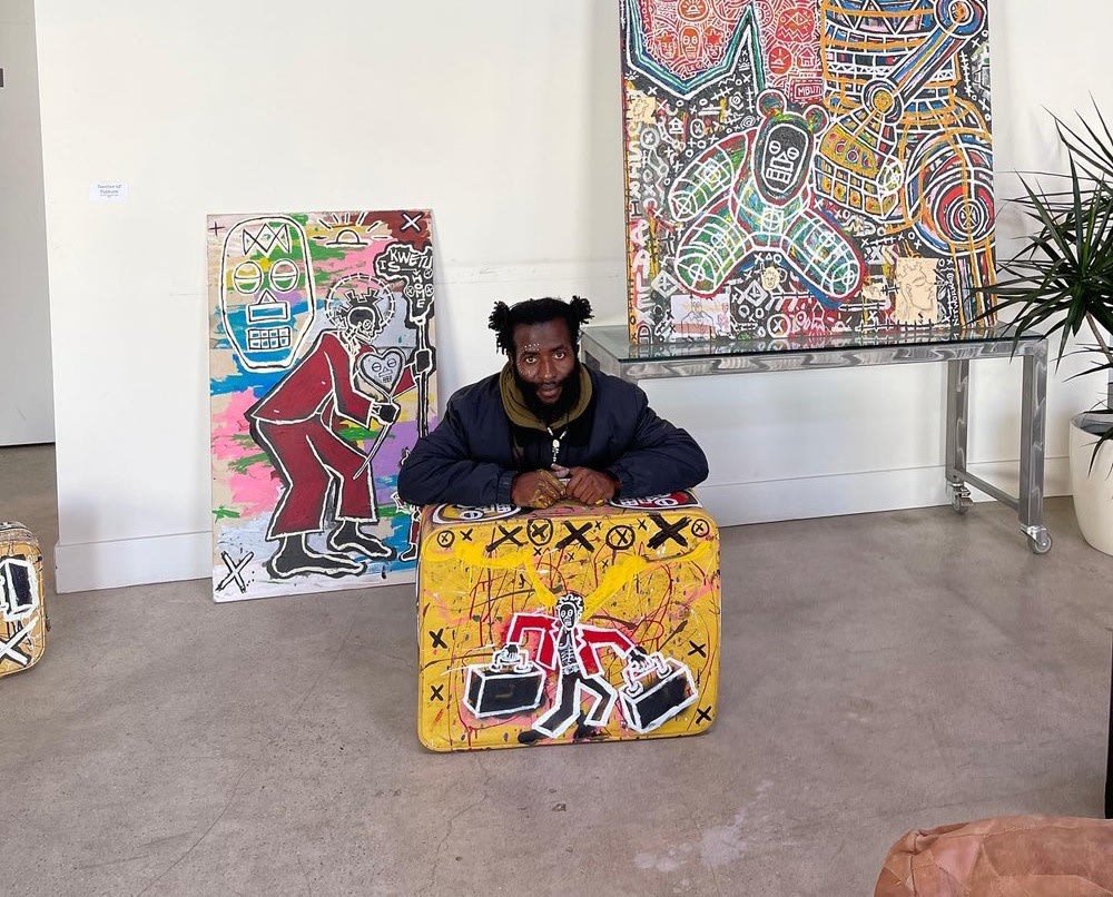 Ba simba is kneeling behind a suitcase he used to create an artwork. He is in a studio with other of his works hanging on the wall and on the floor