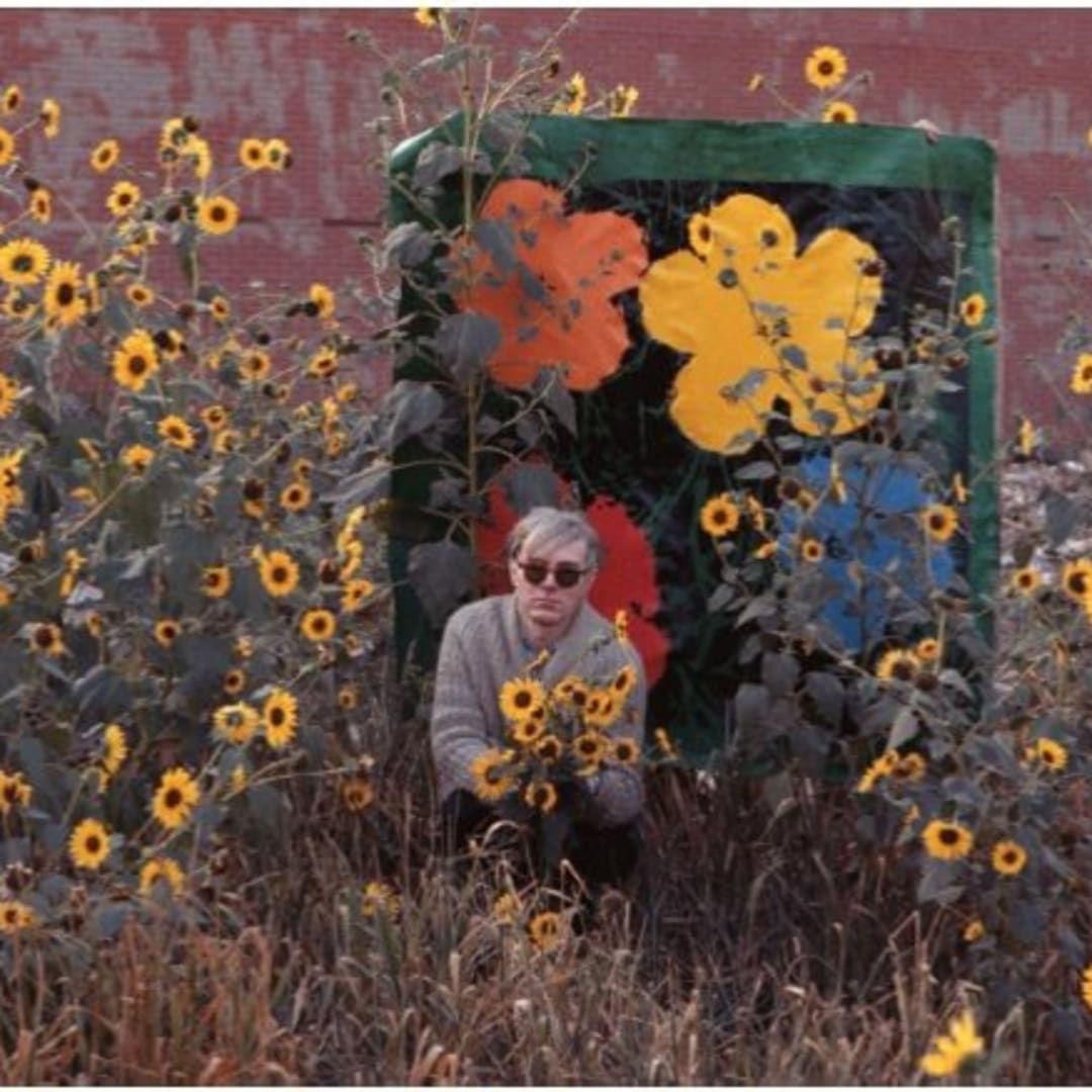 Andy Warhol surrounded by flowers in front of Flower with an early Flowers canvas, Queens, New York, 1964. ©2010 William John Kennedy, ©2012 Andy Warhol Foundation / Artists Rights Society (ARS), New York.