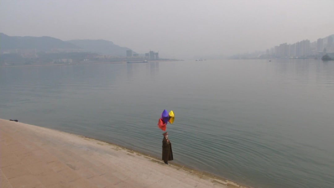 A video still from the Video No.3 out of 7 of Chen Qiulin's The Empty City created in 2012; the still shows the artist Chen Qiulin in a dark green military coat stands on the shore holding balloons of various colors.