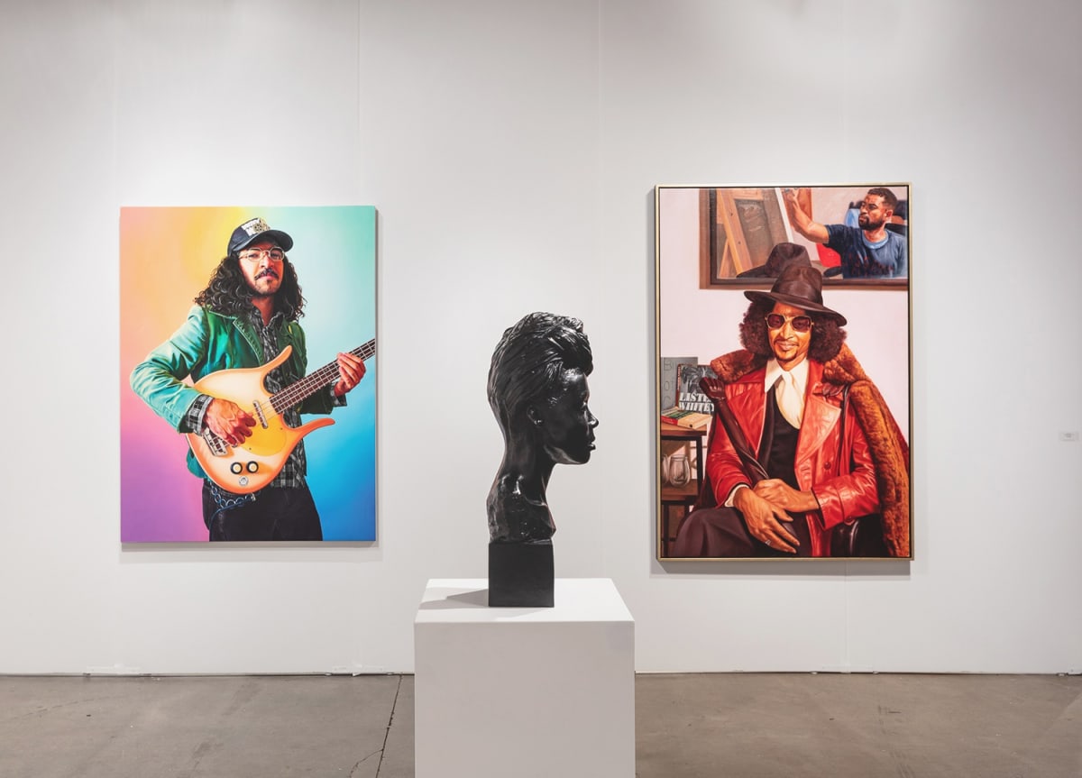 Installation view of an art booth at Expo Chicago art fair. There are two large figurative paintings of men and a sculpture of a bust of a woman sitting on a base in the center.