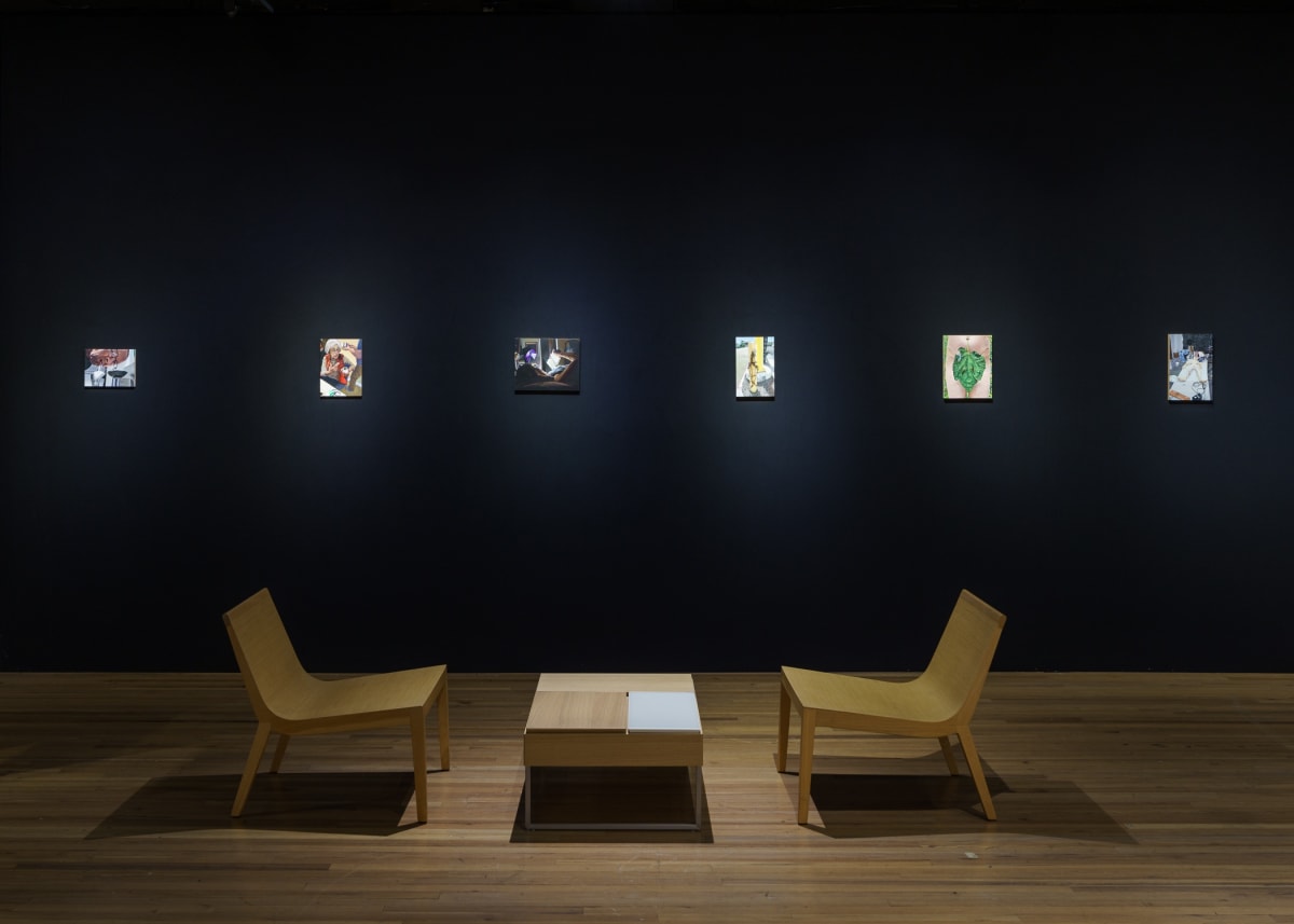 Small oil paintings in an art fair booth against a black wall illuminated by spotlight and two chairs.
