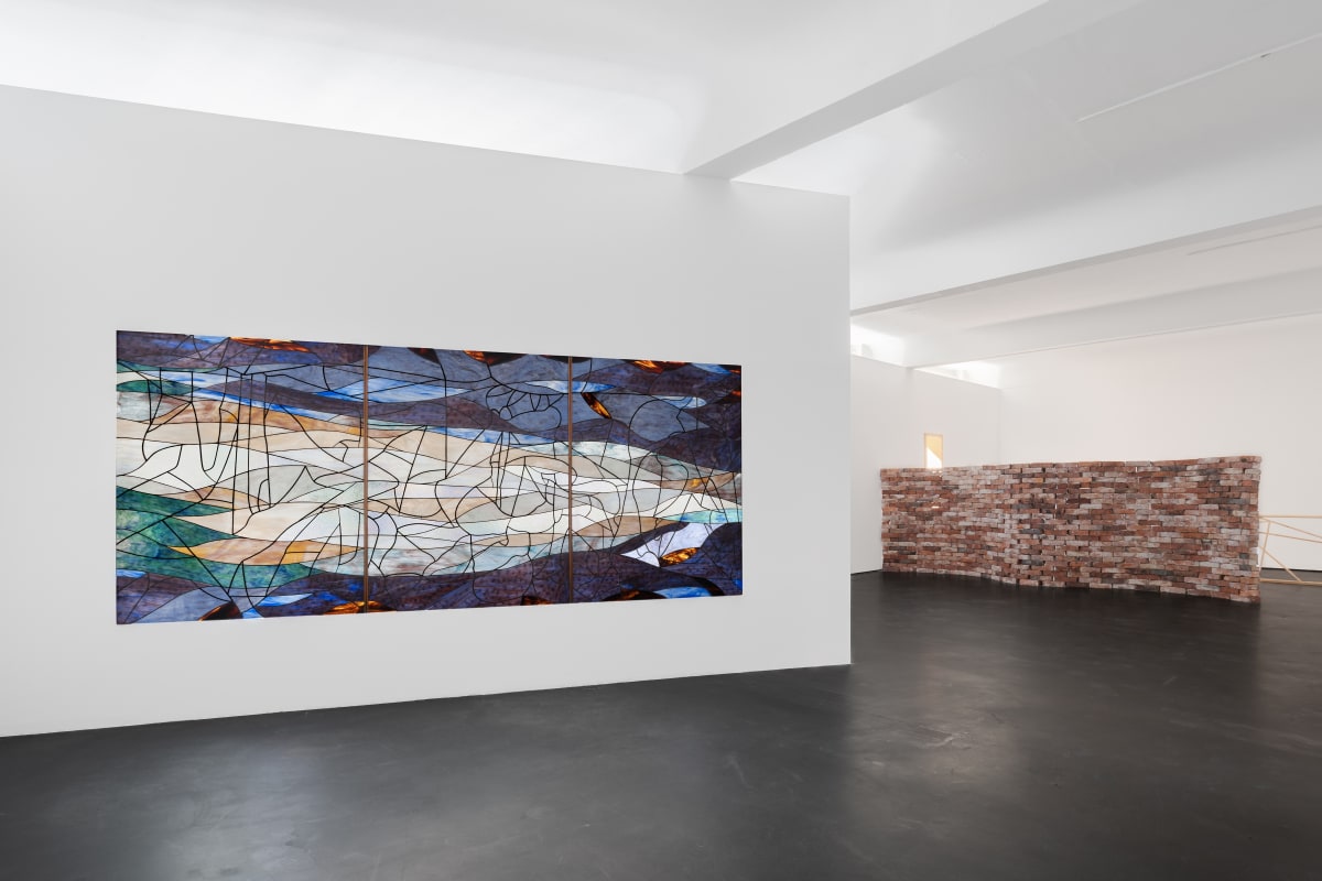 Installation view five | rindon johnson | Photo: Marjorie Brunet Plaza. White room with a dark blue, green and orange stained glass window made up of 3 panels on a plain wall, a red-brown brick wall behind it and a dark floor.