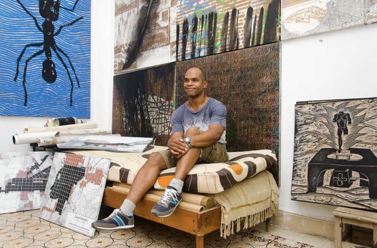 A bald dark-skinned man sits holding his knee on a bed surrounded by artworks.
