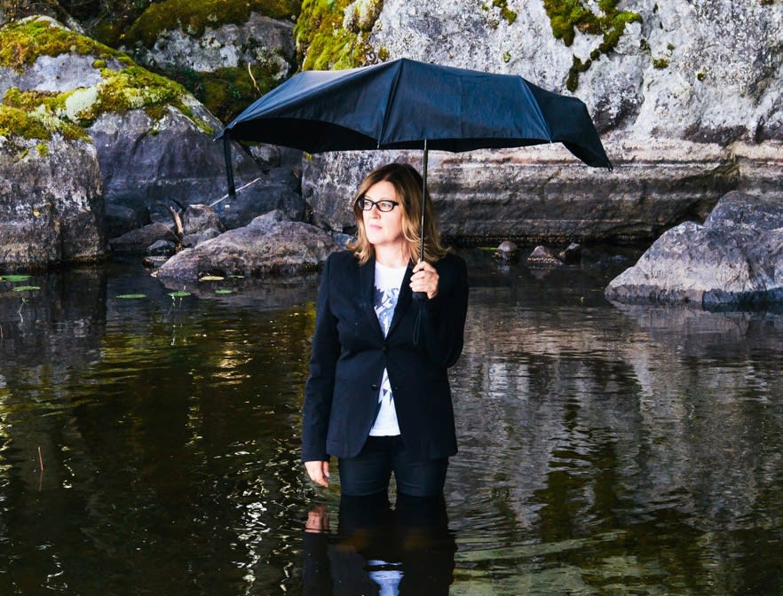 A woman with an umbrella in a lake surrounded by moss and rocks. 