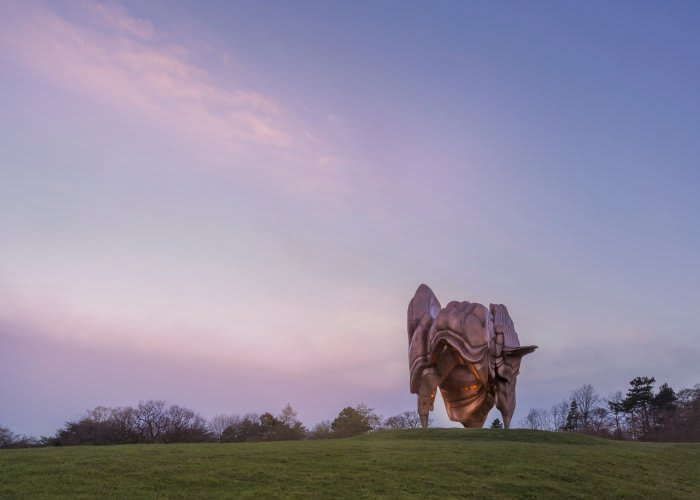 An amorphous large scale bronze sculpture sits in a field. The sky is pink, indicating sunrise or sunset.