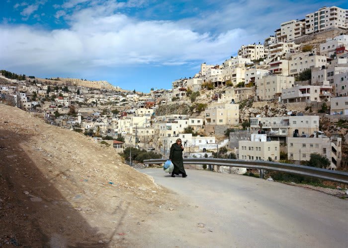 Image of a cloaked figure walking up a dusty road set against a urban desert city.