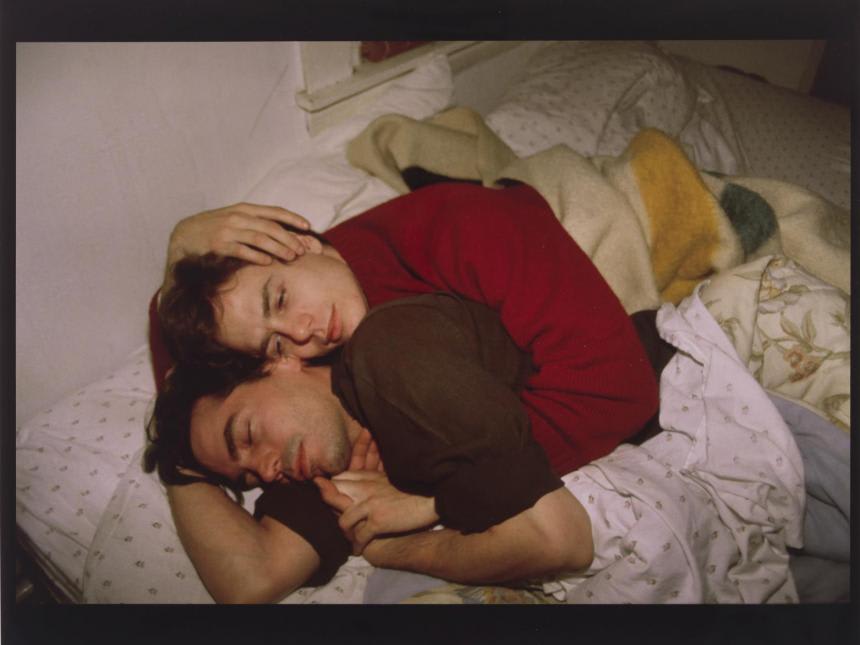 Image of two men wearing sweaters embracing in bed. 