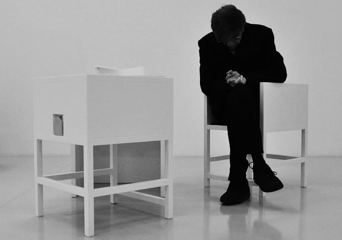 Black and white image of person sitting in geometric white chair. They bow their head and clasp their hands, almost in prayer.