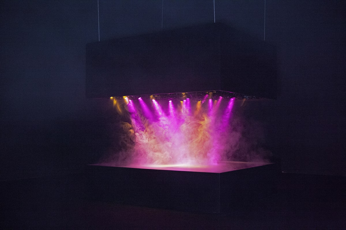 A dark room is illuminated by colored steam between two large stone slabs, one hanging and one on the ground.
