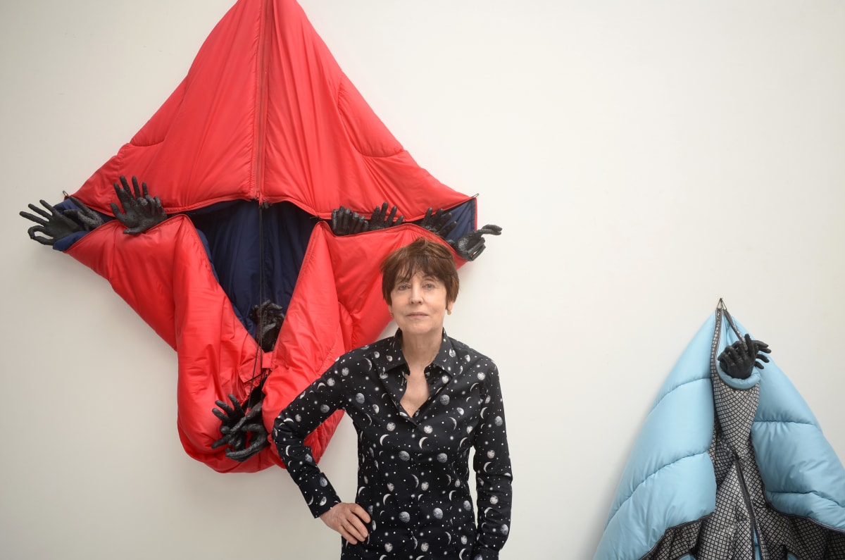 A woman stands in front of a red sleeping bag with multiple casts of hands sticking out, hanging on the wall.