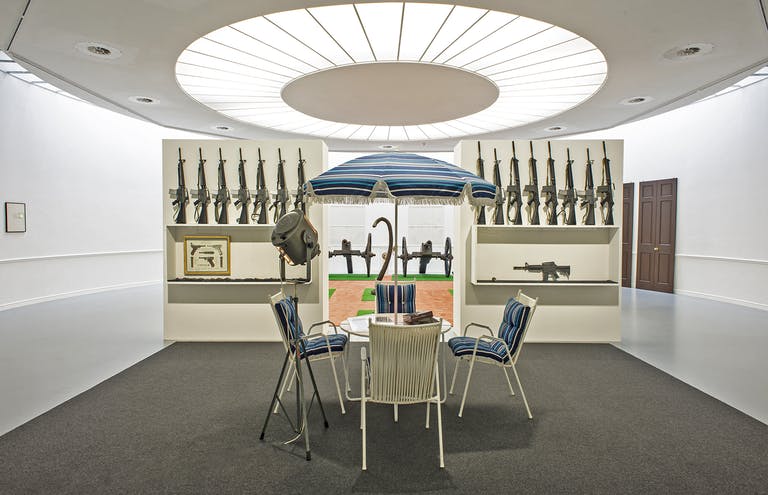 A rendering of a table, umbrella and chairs with cushions in front of a rack of machine guns under a circular skylight.