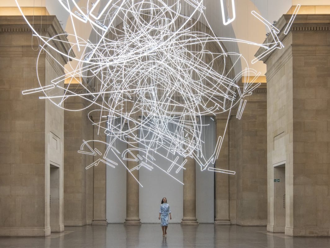 A large marble room features a hanging neon sculpture composed of long, thin and bending lines. 