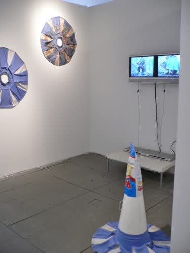 Brock Enright. Wall (left to right): Skirt 1A (FOREST), 2006. Cheerleader skirt, 32 inches in diameter. Skirt 1B (FOREST), 2006. Cheerleader skirt, tomato sauce. 32 inches in diameter. Untitled (Forest), 2006. Dual monitor video loop, edition 1/2, 2:37 min. Floor: Ruffles (FOREST), 2006. Cheerleader skirt, megaphone, and Ruffles bag. 36 x 32 x 32 inches.
