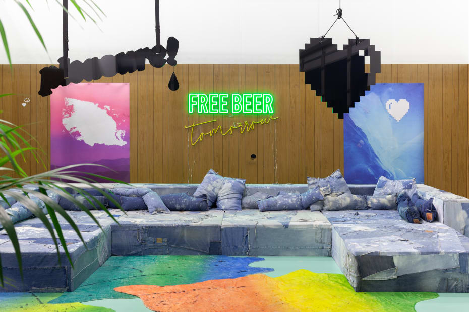 Wendy White. Free Beer Tomorrow, 2019. Hand- carved wood paneling, repurposed distressed denim, upholstery foam and wood custom furniture, digital prints on neoprene, hand painted faux calfskin rugs, paper cigarettes, custom floor paint, LED sign, live plants, Dimensions variable