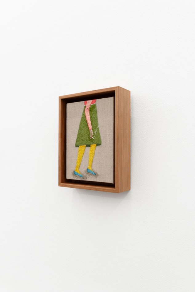 Embroidered figure holding a cigarette, wearing a knee length green skirt and a pink long sleeve top with blue flat shoes.
