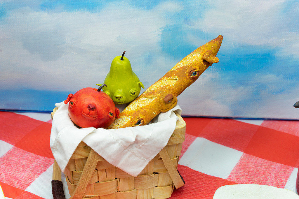 Installation featuring two bottles of wine with dog faces, two sandwiches with dog faces, a picnic basket with a baguette, an apple and a pear all with dog faces on top of a red and white checkered table clothe and a painted sky with clouds in the shape of dogs.