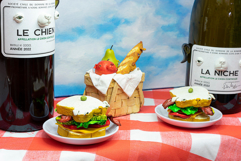 Installation featuring two bottles of wine with dog faces, two sandwiches with dog faces, a picnic basket with a baguette, an apple and a pear all with dog faces on top of a red and white checkered table clothe and a painted sky with clouds in the shape of dogs.