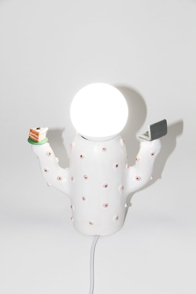 back of Jen Dwyer lamp sculpture of white cactus with pink lips holding laptop and slice of cake. Lightbulb sits on top