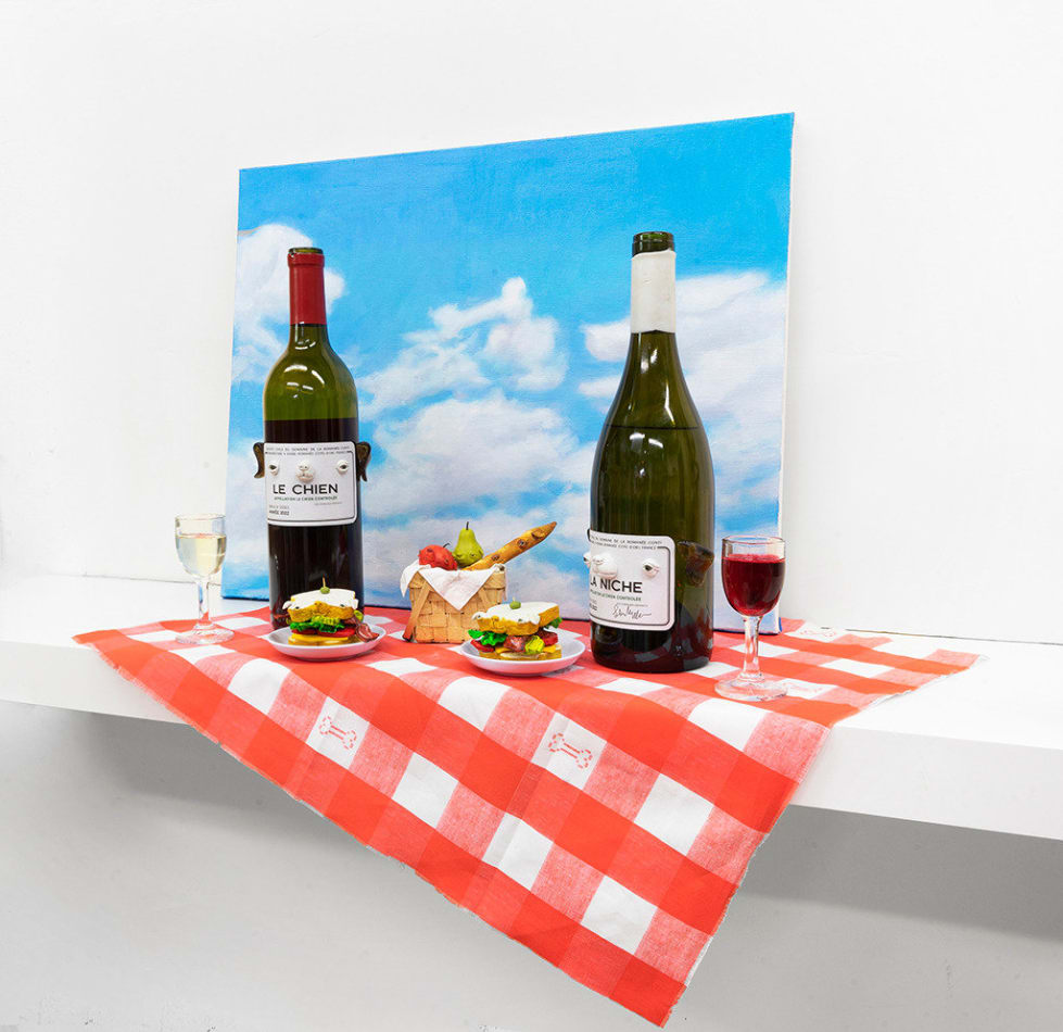 Installation featuring two bottles of wine with dog faces, two sandwiches with dog faces, a picnic basket with a baguette, an apple and a pear all with dog faces on top of a red and white checkered table clothe and a painted sky with clouds in the shape of dogs.