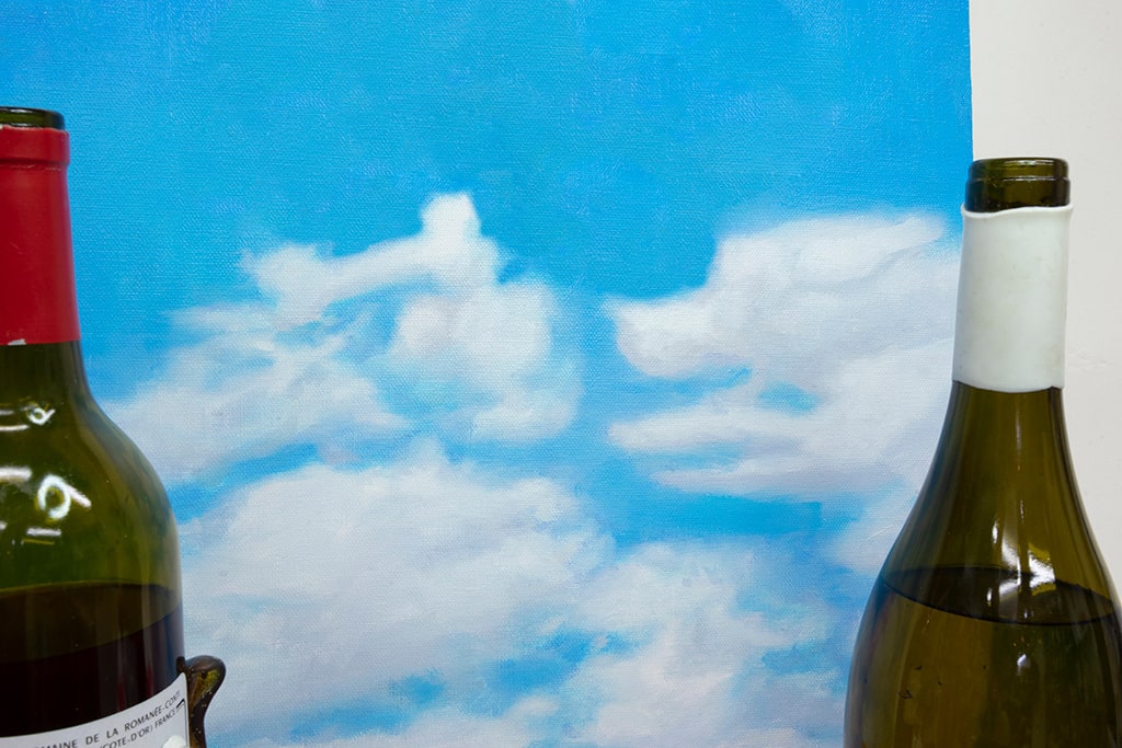 Installation featuring two bottles of wine with dog faces, two sandwiches with dog faces, a picnic basket with a baguette, an apple and a pear all with dog faces on top of a red and white checkered table clothe and a painted sky with clouds in the shape of dogs.