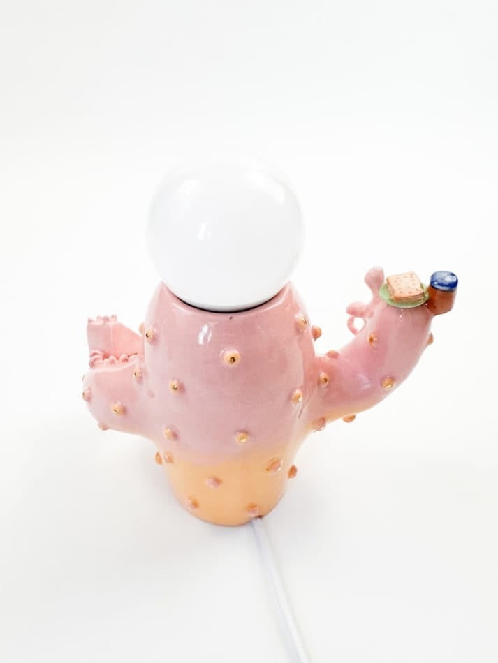 Lamp sculpture of a cactus like object with lips and two arms holding a plate with a sandwich and the other a bag of chips