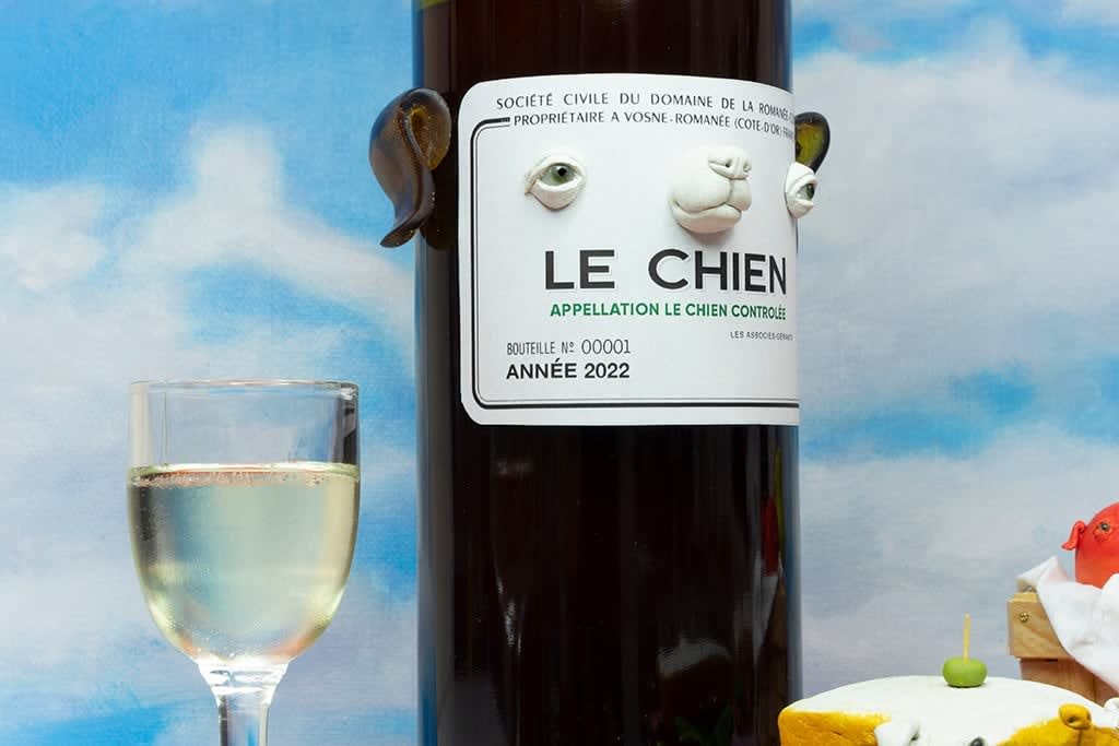 Installation featuring two bottles of wine with dog faces, two sandwiches with dog faces, a picnic basket with a baguette, an apple and a pear all with dog faces on top of a red and white checkered table clothe and a painted sky with clouds in the shape of dogs.