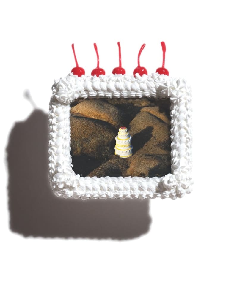A picture frame that looks like a white cake with 5 cherries on top. Inside of the frame is a picture of a white 3 layered cake with cherries on top in the middle of a bunch of boulders.