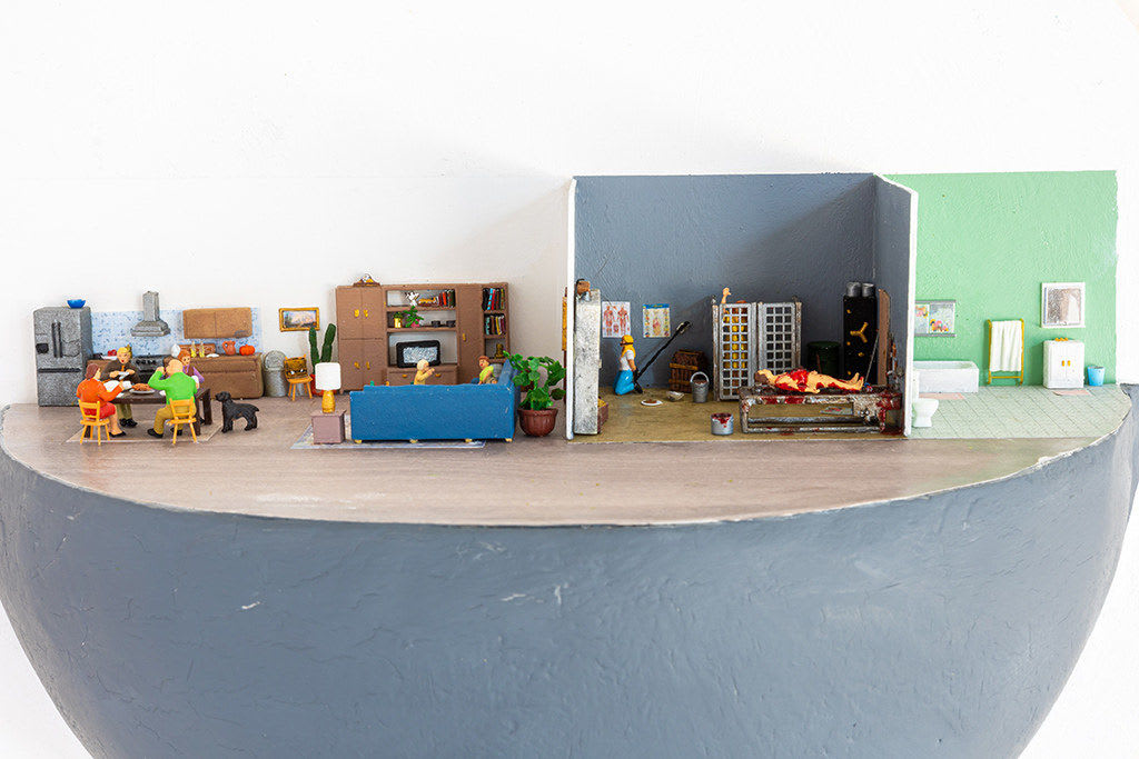 miniature diorama by Abigail Goldman of the interior of house with three rooms, one in which a family is eating dinner and another with people chained and bleeding