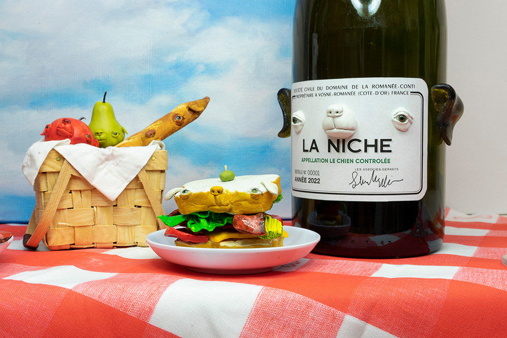 Installation featuring two bottles of wine with dog faces, two sandwiches with dog faces, a picnic basket with a baguette, an apple and a pear all with dog faces on top of a red and white checkered table clothe and a painted sky with clouds in the shape of dogs.