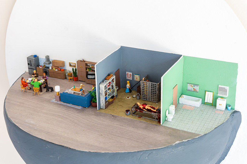 miniature diorama by Abigail Goldman of the interior of house with three rooms, one in which a family is eating dinner and another with people chained and bleeding