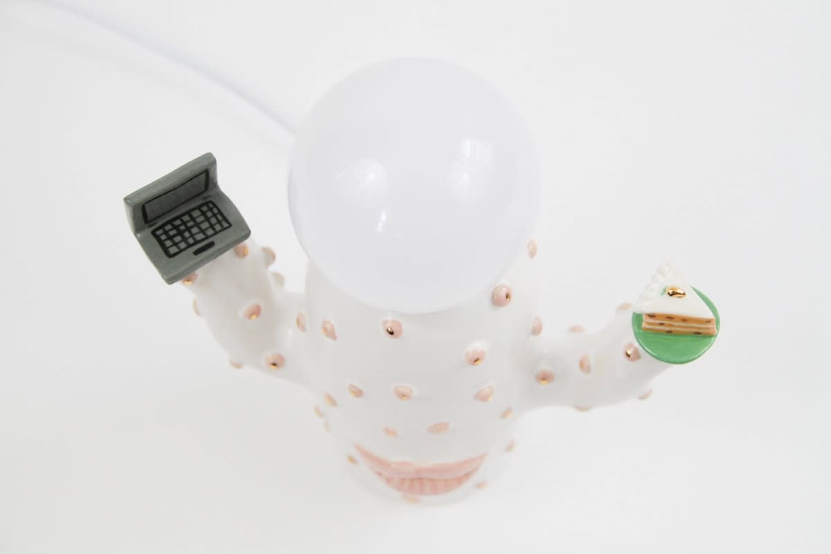 aerial view of Jen Dwyer lamp sculpture of white cactus with pink lips holding laptop and slice of cake. Lightbulb sits on top