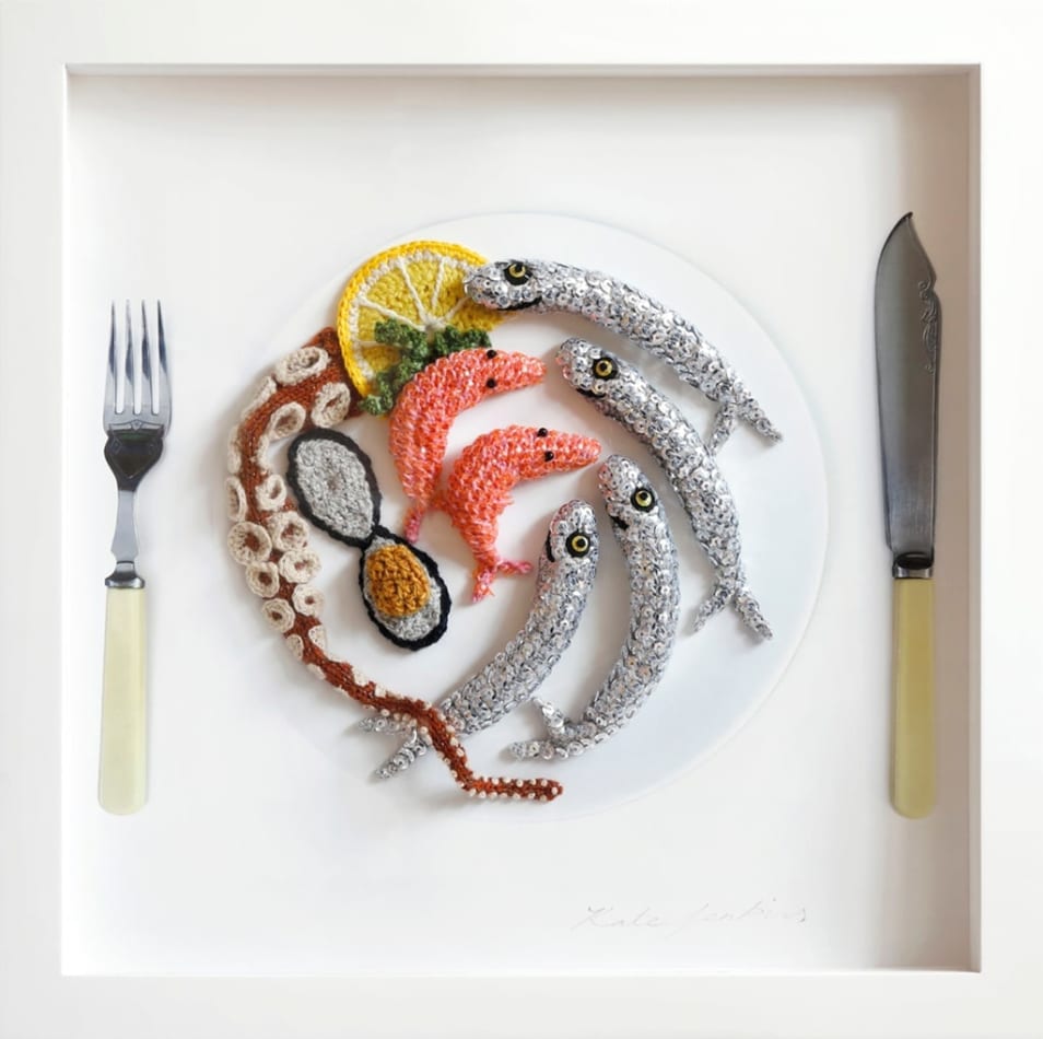Soft sculpture of crocheted sardines, shrimp and octopus on a white place with a lemon and fork and knife