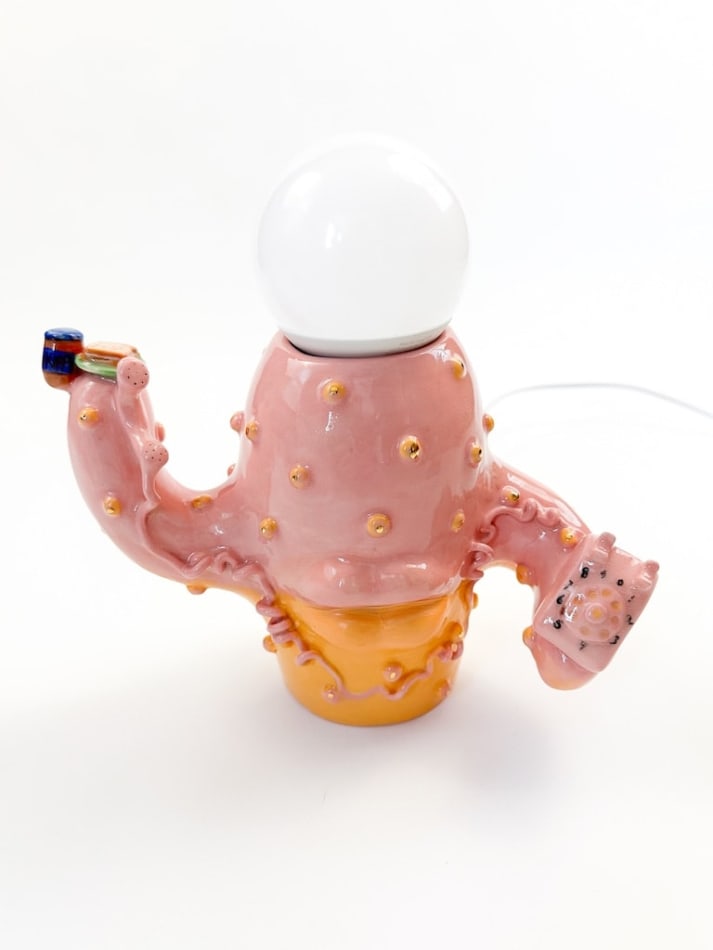 Lamp sculpture of a cactus like object with lips and two arms holding a plate with a sandwich and the other a bag of chips