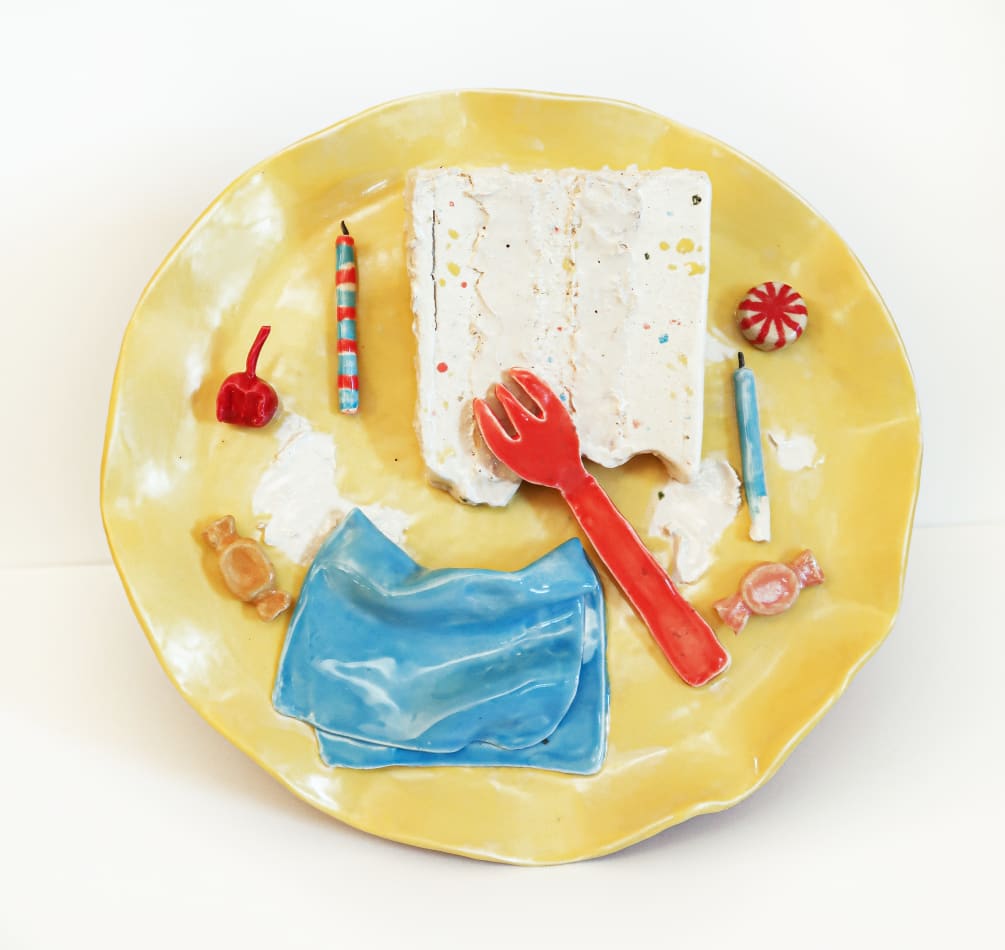 A ceramic plate with ceramic fork, piece of cake, candies, birthday candles and a cherry on it.