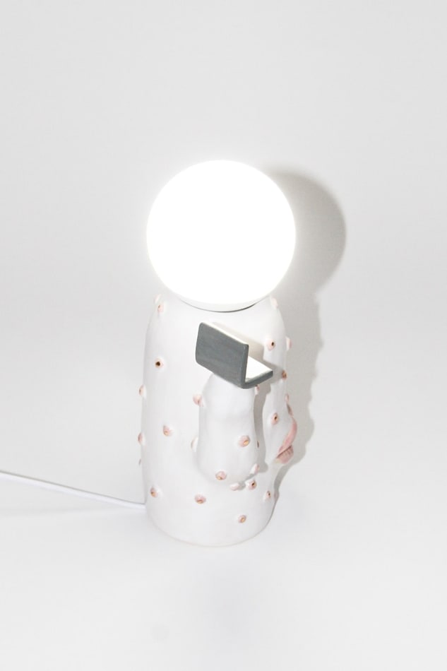 side of Jen Dwyer lamp sculpture of white cactus with pink lips holding laptop and slice of cake. Lightbulb sits on top