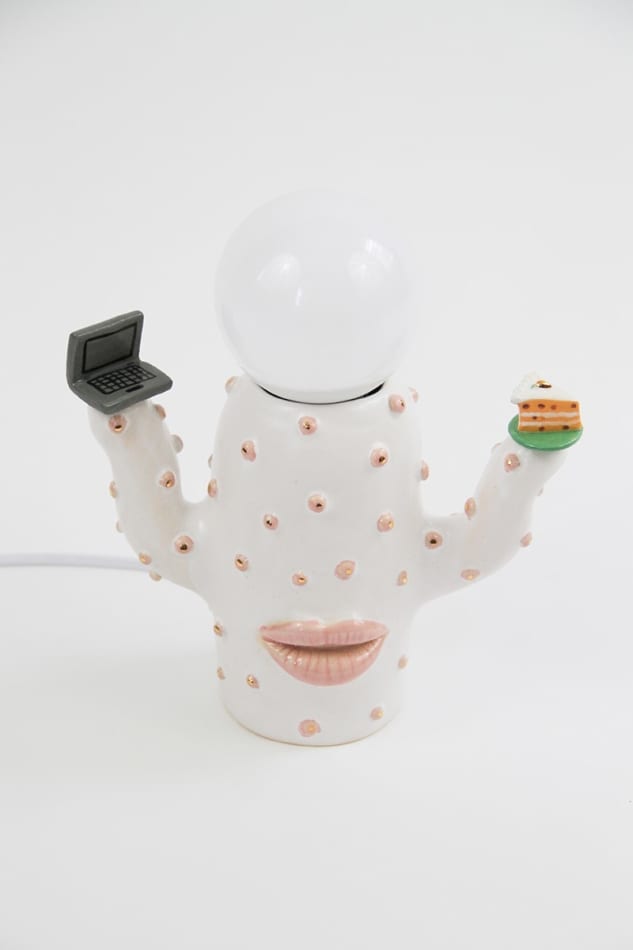 Jen Dwyer lamp sculpture of white cactus with pink lips holding laptop and slice of cake. Lightbulb sits on top