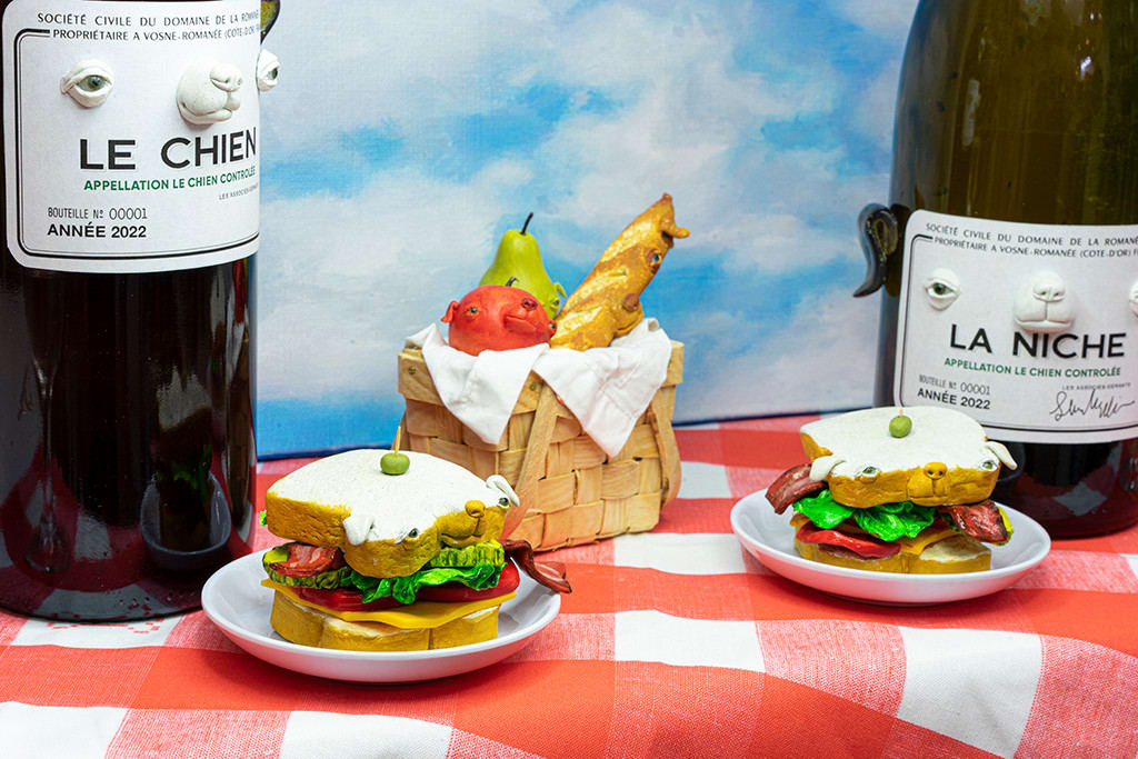 Installation featuring two bottles of wine with dog faces, two sandwiches with dog faces, a picnic basket with a baguette, an apple and a pear all with dog faces on top of a red and white checkered table clothe and a painted sky with clouds in the shape of dogs.