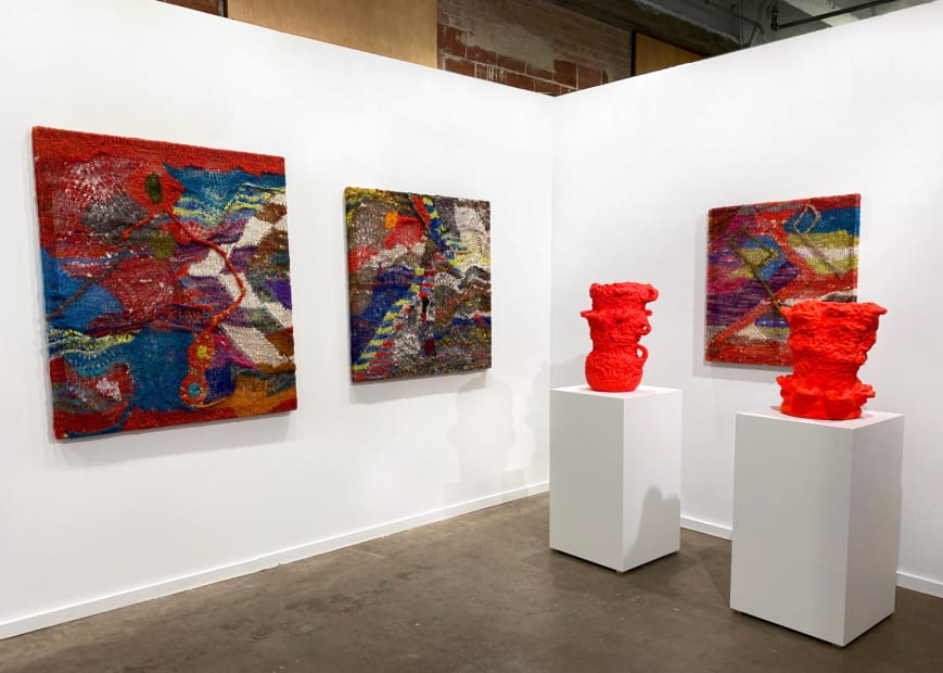 Installation view at the Dallas Art Fair with several colorful knitted works by Channing Hansen and two red pots on plinths by Beverly Semmes