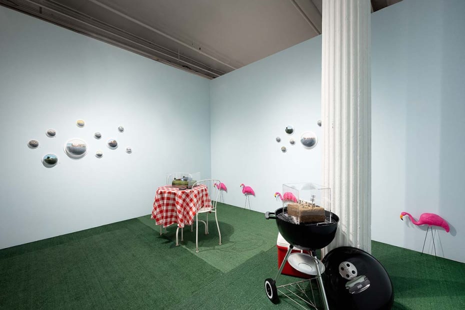 Abigail Goldman installation at Outsider Art Fair. Picturesque suburban backyard with white picket fence in forground, red checkered tablecloth bistro table and grill / cooler visible. Abigail's work hangs in clusters on walls and on top of grill and tabl