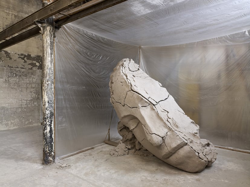 image of Mark Manders installation with split clay head on table and large dry clay head with plastic