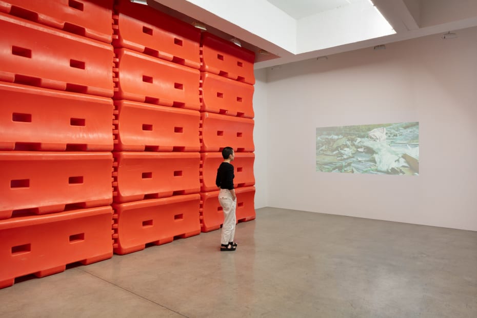 Installation image, Karyn Olivier, How A Home Is Made, a figure stands in front of a plastic orange barricade stacked to ceiling and a projection on the right