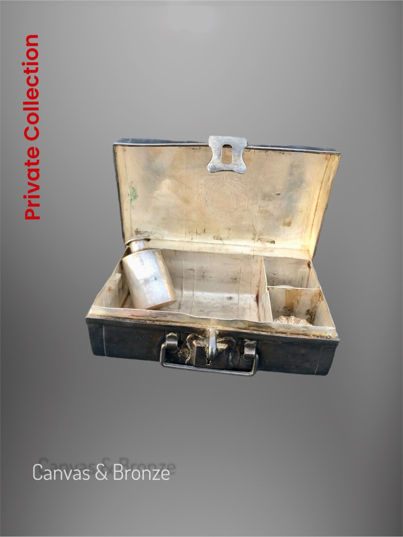 Open vintage metal toolbox with compartments, containing a small silver flask inside, against a gray background.